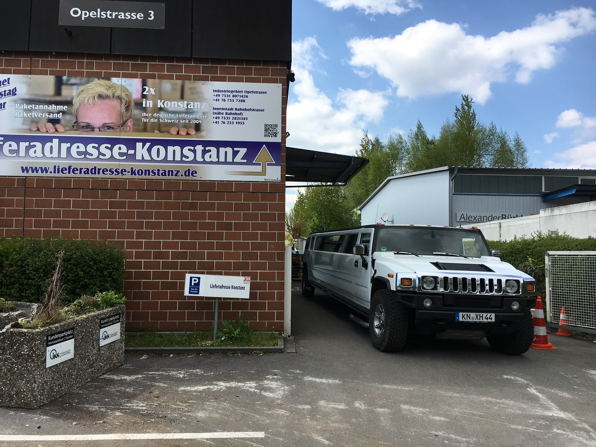 Weiße Hummer H2 Stretchlimousine vor der Einfahrt der Lieferadresse Konstanz in der Opelstraße 3 mit Firmenschild und Kundenparkplätzen